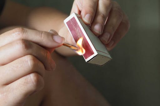 The hands of a woman are pictured striking a match against the side of a matchbox. There is a flame starting.